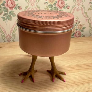May include: A copper-colored jar with a lid, resting on two chicken feet with pink painted nails. The lid is inscribed with the word "FELICITY" in a decorative font. The jar is sitting on a wooden surface.