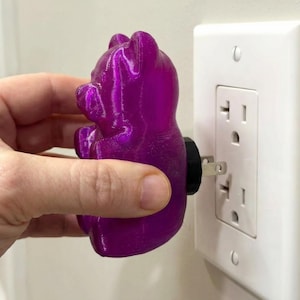 Hand shown in the process of plugging a translucent purple gummy bear night light into a standard white wall outlet. This close-up view highlights the tactile details of the retro kitsch glowing candy art and the convenience of this funky home accent plug in luminary. Perfect for those looking to buy an illuminated bear night light, this unique plug in outlet light instantly transforms boring spaces with a brilliant weirdcore room vibe and functions as the ultimate dopamine decor apartment gift.