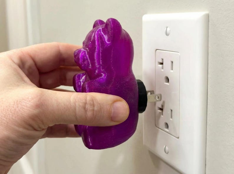 Hand shown in the process of plugging a translucent purple gummy bear night light into a standard white wall outlet. This close-up view highlights the tactile details of the retro kitsch glowing candy art and the convenience of this funky home accent plug in luminary. Perfect for those looking to buy an illuminated bear night light, this unique plug in outlet light instantly transforms boring spaces with a brilliant weirdcore room vibe and functions as the ultimate dopamine decor apartment gift.