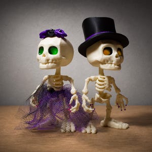 May include: Two off-white skeleton figurines standing back-to-back. The left skeleton wears a purple floral headpiece and a purple and black tulle skirt. The right skeleton wears a black top hat with a purple band. Both have colored eye sockets.