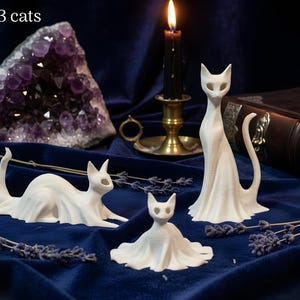 May include: A set of three white cat figurines in various poses. One cat is seated, another is lying down, and the third is standing. The text "Set of 3 cats" is in the upper left corner. A lit candle and a large amethyst crystal are in the background.