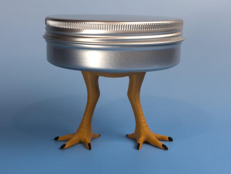 May include: A round, silver-colored metal tin with a screw-top lid, supported by two yellow, textured bird-like legs with black-tipped claws. The container is positioned upright, showcasing a metallic finish and realistic avian-inspired limbs.