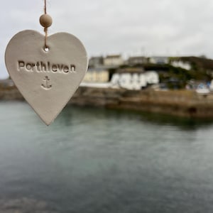 May include: White ceramic heart-shaped ornament with the text "Porthleven" and an anchor symbol. The ornament is hanging from a string with a wooden bead.