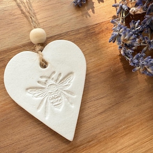 May include: A white heart-shaped clay ornament featuring an embossed bee design on the front. The ornament is suspended by a piece of natural jute twine and includes a single round wooden bead above the heart-shaped piece.