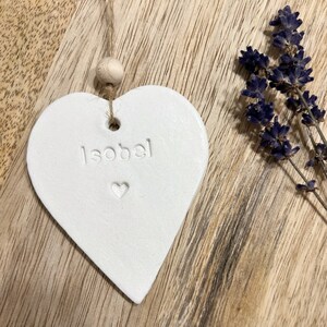 May include: White ceramic heart ornament with the name "Isobel" inscribed on it. The heart is hanging from a twine string with a wooden bead.