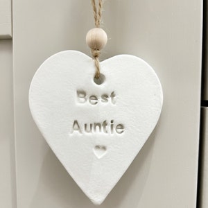 May include: White ceramic heart-shaped ornament with a twine hanger and a wooden bead. The ornament says "Best Auntie" with a small heart symbol.