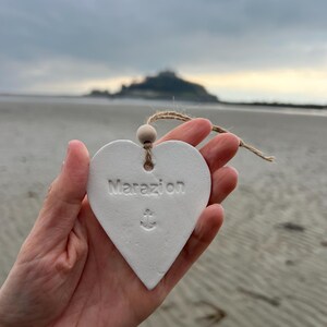 May include: White heart-shaped ceramic ornament with the word "Marazion" and an anchor symbol. The ornament is being held in a hand with a blurry background of a beach and a hill in the distance.