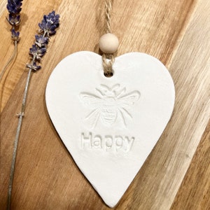 May include: White ceramic heart-shaped ornament with a bee design and the word "Happy" embossed on it. The ornament is hanging from a twine string with a wooden bead at the top.