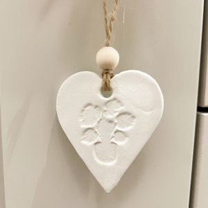 May include: White ceramic heart ornament with a raised design of leaves and a small circle in the centre. The ornament is hanging from a twine string with a wooden bead.