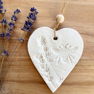 May include: White ceramic heart-shaped ornament with a raised design of lavender flowers and a bee. The ornament has a twine hanger with a wooden bead.