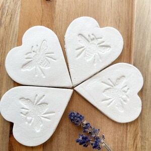 May include: Four white ceramic heart-shaped ornaments with a raised bee design. The ornaments are arranged in a circle with a sprig of lavender in the centre.
