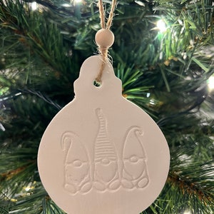 May include: A white, round Christmas ornament featuring three embossed gnome designs. It is suspended by a natural twine string and a wooden bead. The ornament hangs against a backdrop of a green Christmas tree.
