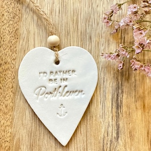 May include: A white heart-shaped hanging ornament made of clay, featuring the embossed text 'I'D RATHER BE IN Porthleven' and a small anchor symbol. The ornament is suspended by a tan jute twine string with a single round wooden bead.