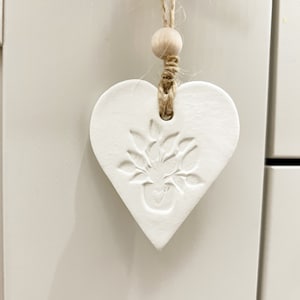 May include: White ceramic heart-shaped ornament with a plant design, hanging from a twine string with a wooden bead.