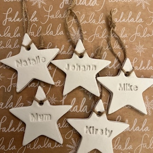 May include: Five white star-shaped Christmas ornaments, each with a name: Natalie, Johann, Mike, Mum, and Kirsty. They have twine loops for hanging. The background is brown wrapping paper with a repeating "Falala" pattern and snowflakes.