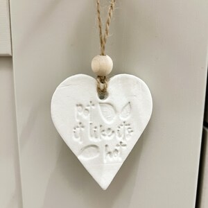 May include: White ceramic heart-shaped ornament with the text "put a little love in hot" hanging from a twine string with a wooden bead.