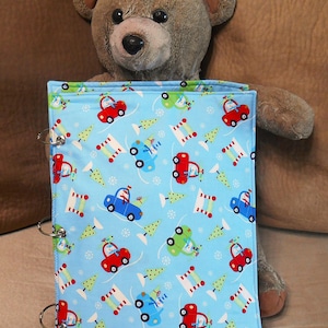 May include: A light blue fabric book cover with a repeating pattern of colorful cars, Christmas trees, and snow. The cover is held open by silver binder rings. A brown teddy bear is behind the book.