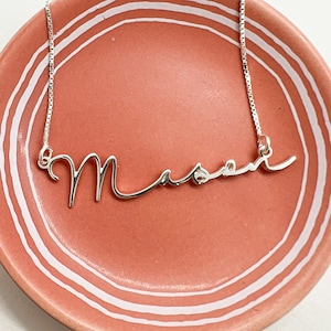 May include: A silver-colored necklace with the name "Mason" in a cursive font. The necklace is displayed on a round, coral-colored dish with white concentric lines.