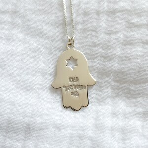 May include: Silver-toned pendant necklace featuring a Hamsa hand design with a Star of David cutout. The pendant has Hebrew text. The necklace hangs on a delicate chain, set against a white fabric background.