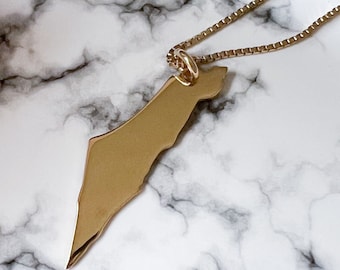 Op de afbeelding: Een goudkleurige hangerketting met de omtrek van een land. De hanger is bevestigd aan een fijne gouden ketting. De ketting wordt gepresenteerd op een wit en grijs marmeren oppervlak.