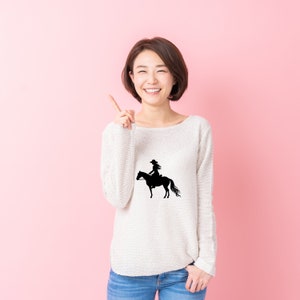 May include: A woman wearing a white sweater with a black silhouette of a cowgirl riding a horse on the front. She is smiling and pointing to her right.
