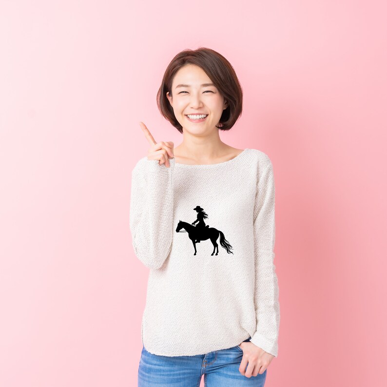 May include: A woman wearing a white sweater with a black silhouette of a cowgirl riding a horse on the front. She is smiling and pointing to her right.