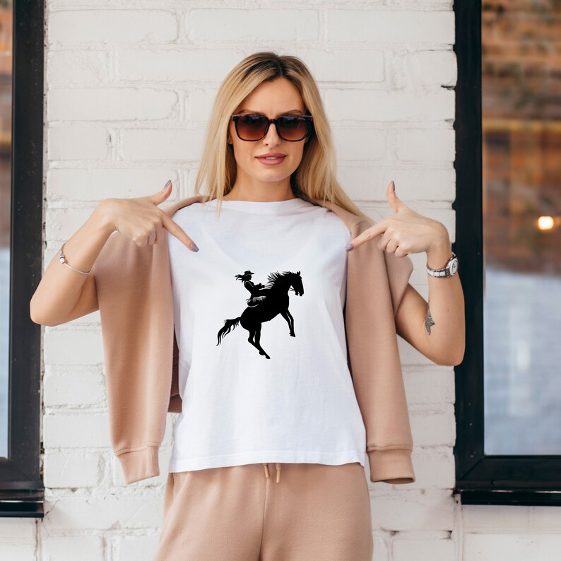 May include: White t-shirt with a black silhouette of a person riding a horse.