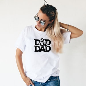 May include: White t-shirt with the black text "D&D DAD". The shirt has short sleeves and a crew neck. The graphic features a stylized dragon and dice.