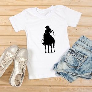 May include: A white t-shirt with a black silhouette of a woman riding a horse. The woman is wearing a hat and holding the reins. The t-shirt is lying on a wooden surface with a pair of white shoes and a pair of blue denim shorts.