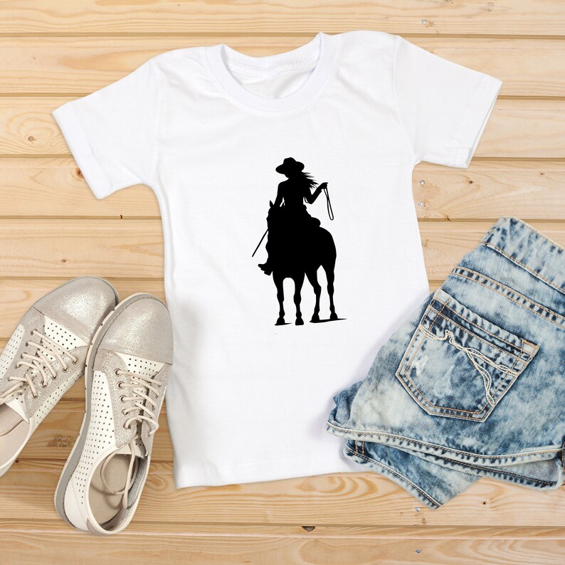 May include: A white t-shirt with a black silhouette of a woman riding a horse. The woman is wearing a hat and holding the reins. The t-shirt is lying on a wooden surface with a pair of white shoes and a pair of blue denim shorts.