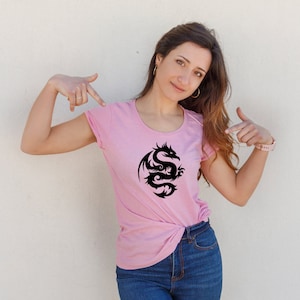 May include: A light pink t-shirt featuring a black dragon design. The dragon is stylized with intricate curves and details. The shirt has a round neckline and short sleeves.
