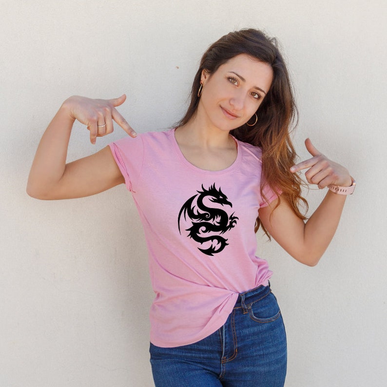 May include: A light pink t-shirt featuring a black dragon design. The dragon is stylized with intricate curves and details. The shirt has a round neckline and short sleeves.