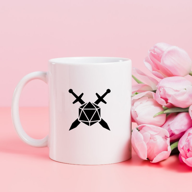 May include: White ceramic mug featuring a black graphic of a 20-sided die crossed with two swords. The mug has a curved handle and is set against a pink background with pink tulips.