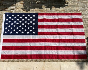 May include: A red, white, and blue American flag with 50 white stars on a blue field. The flag is hanging on a brick wall.