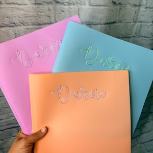 May include: Three plastic folders in pastel pink, light blue, and peach. Each folder features the name Darcie embroidered in a cursive font using small, sparkling rhinestones. The folders are stacked, showing the front covers clearly.