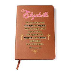 May include: A brown leather journal with the name "Elizabeth" embossed in pink lettering. The journal features a gold inscription with a bible verse: "She is clothed with Strength and Dignity: She can laugh at the days to come. She speaks with Wisdom, and Faithful instruction is on her tongue. Proverbs 31"