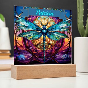 May include: A stained glass style image of a dragonfly with a purple, blue, and yellow background. The image is on a wooden stand and has the name "Patricia" written on a blue banner at the top.