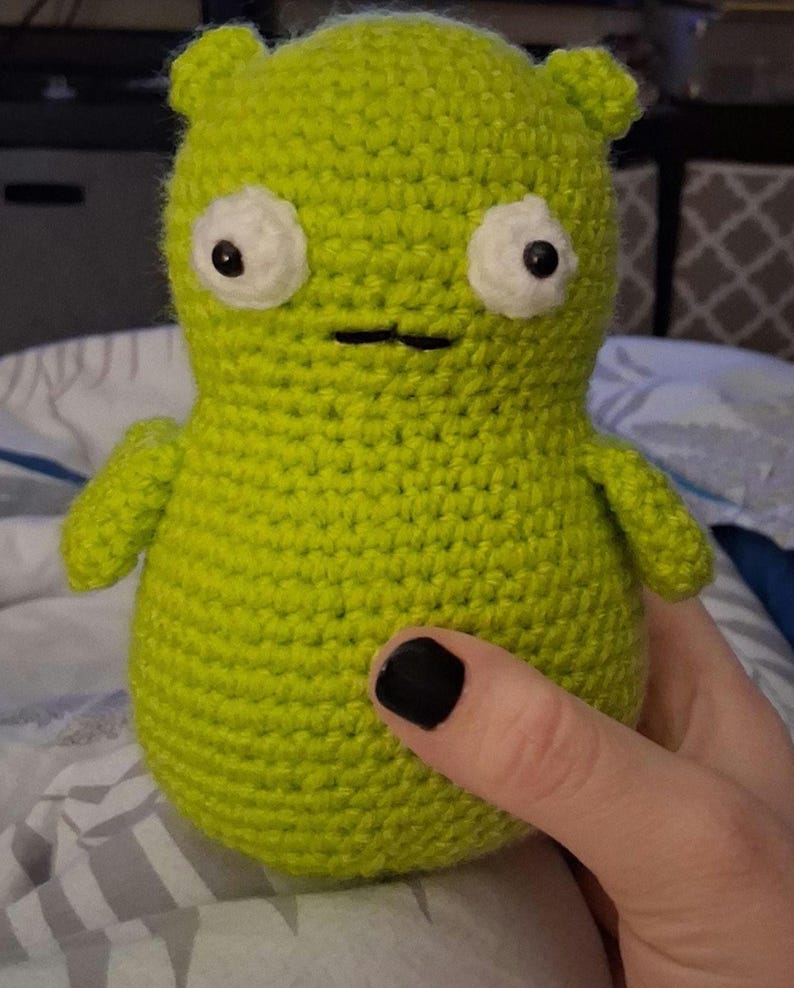 K&ouml;nnte beinhalten: Eine handgefertigte, limettengr&uuml;ne H&auml;kelpuppe mit wei&szlig;en Augen und schwarzen Pupillen. Die Puppe hat kleine Ohren, einen einfachen Mund und kurze Arme. Die Puppe wird von einer Person mit schwarzem Nagellack gehalten.