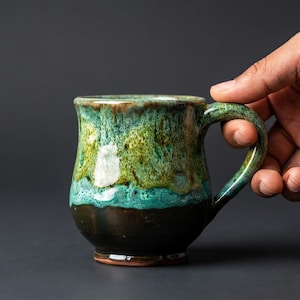 May include: A handcrafted ceramic mug with a unique glaze. The mug features a dark brown base, transitioning to a turquoise and green center, and a brown rim. The mug is held by a hand against a dark gray background.