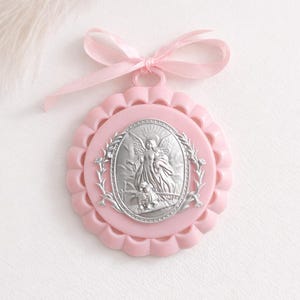 May include: A pink, scalloped-edged ornament with a silver-toned angel illustration. The ornament has a pink ribbon tied in a bow at the top. The angel illustration is surrounded by a silver-toned laurel wreath.