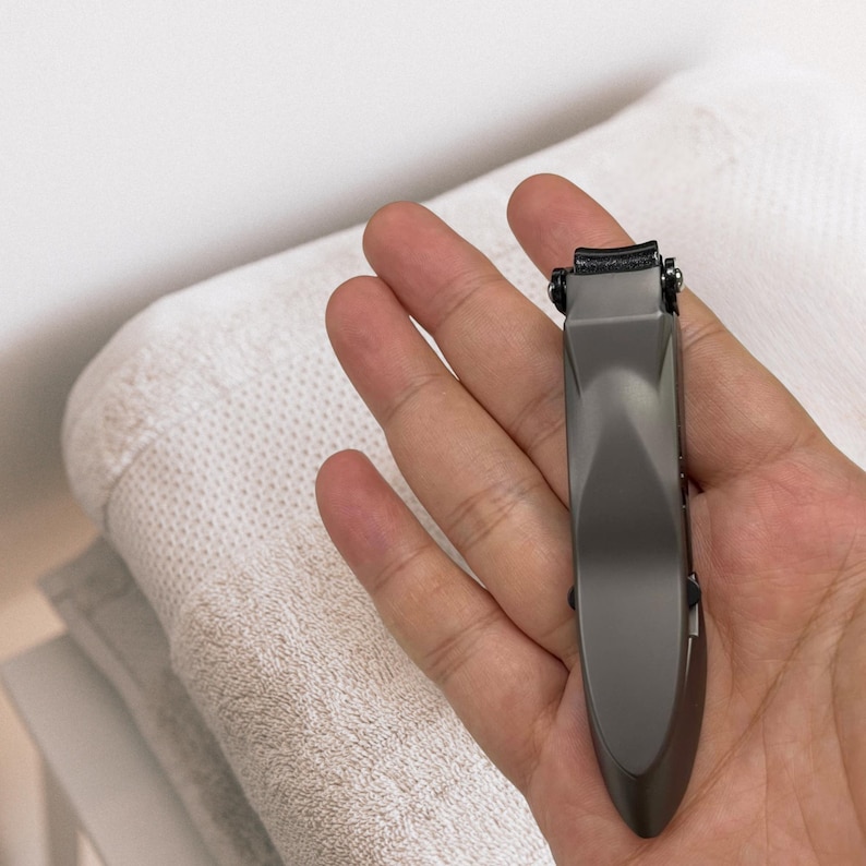 Puede incluir: Un corta&uacute;&ntilde;as gris oscuro con un borde de corte curvo y un mango largo y ergon&oacute;mico. El corta&uacute;&ntilde;as se sostiene en una mano abierta, con una toalla blanca en el fondo.