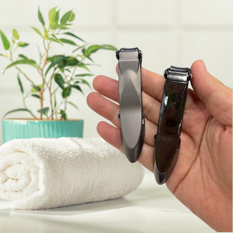 Puede incluir: Dos corta&uacute;&ntilde;as, uno gris y otro negro, se sostienen en una mano. Los corta&uacute;&ntilde;as tienen un dise&ntilde;o elegante y moderno. Una toalla blanca enrollada y una planta en maceta est&aacute;n en el fondo.