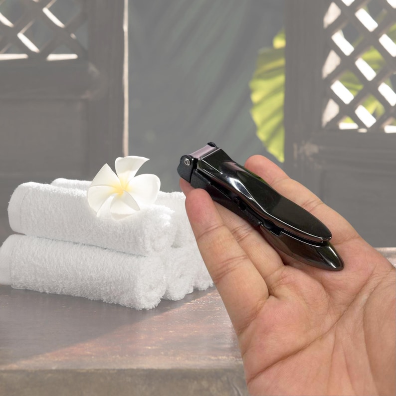 Puede incluir: Un corta&uacute;&ntilde;as negro brillante se sostiene en una mano. Hay toallas blancas y una flor blanca en el fondo. La herramienta est&aacute; dise&ntilde;ada para el cuidado de las u&ntilde;as.