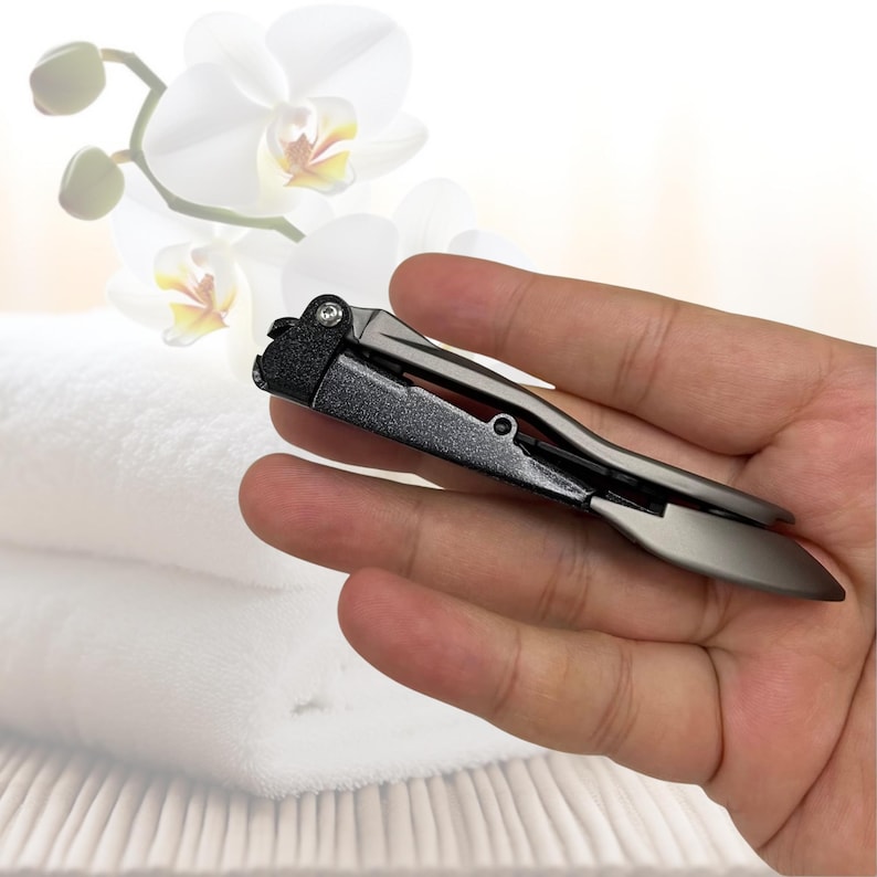 Puede incluir: Un corta&uacute;&ntilde;as gris oscuro con un acabado mate. El corta&uacute;&ntilde;as se sostiene en una mano, mostrando su dise&ntilde;o elegante. El fondo presenta toallas blancas y un arreglo floral borroso.