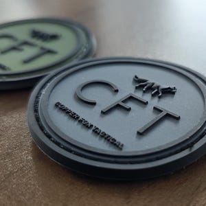 May include: Two round, textured patches. One is dark gray with the letters "CFT" and a fox logo, and the words "COPPER FOX TACTICAL" around the edge. The other patch is olive green with the letters "CFT" and a logo. Both patches have a black border.