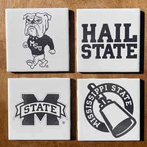 May include: Four square white ceramic coasters with black graphics. One coaster features a bulldog wearing an MSU sweater. Another says "HAIL STATE." The third has a "STATE" logo, and the fourth has a Mississippi State graphic.