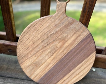May include: Round wooden cutting board with a handle. The board is made of two different types of wood, one light and one dark. The handle has a ring for hanging.