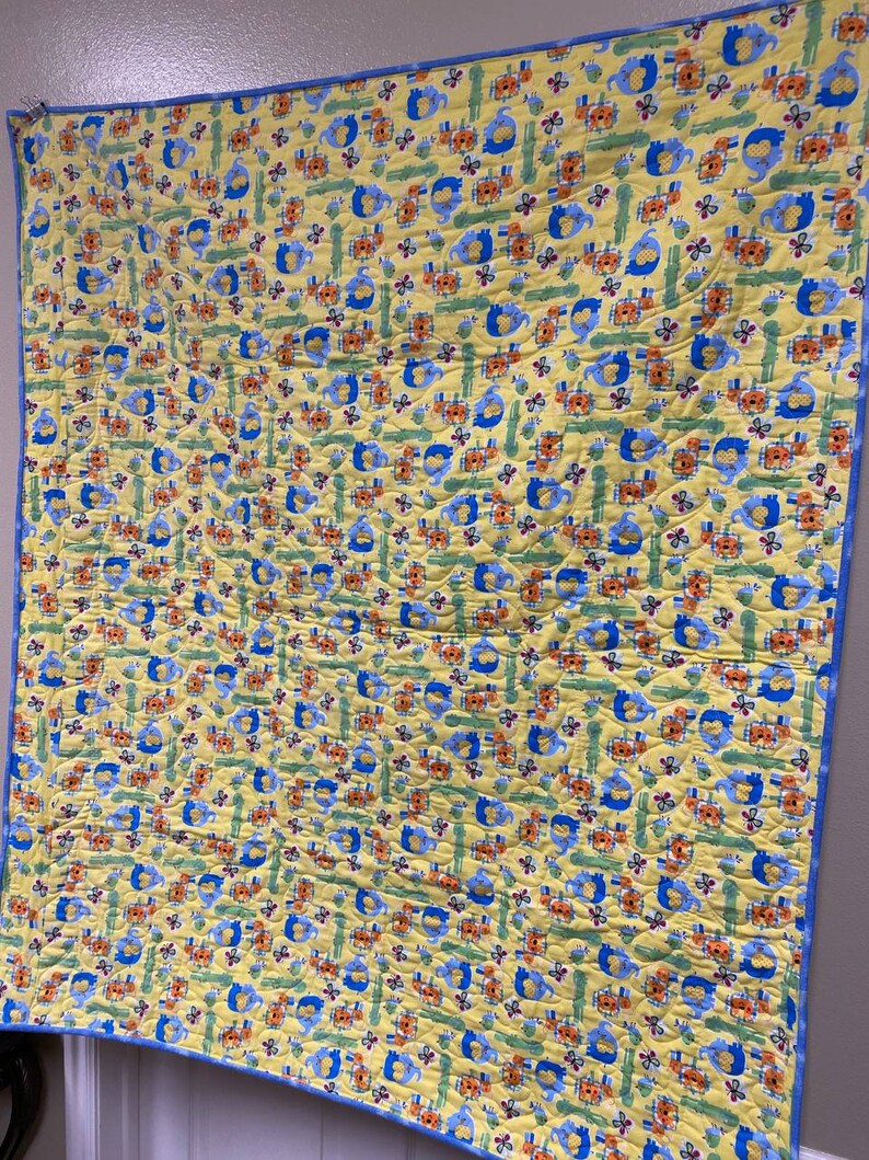 May include: A yellow baby quilt with a blue border. The quilt features a repeating pattern of blue elephants, orange flowers, and green leaves.