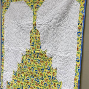 May include: A white quilted baby blanket with a yellow and blue border. The blanket features a quilted design of two large angel wings.