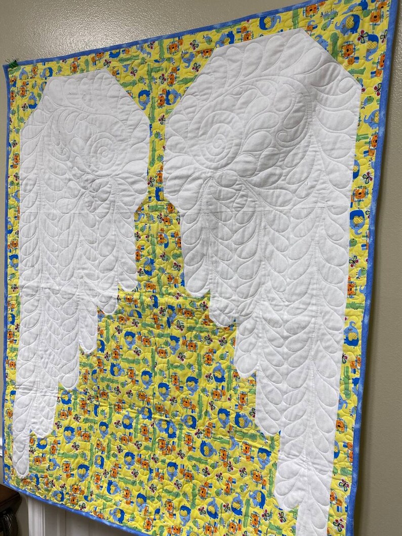 May include: A white quilted baby blanket with a yellow and blue border. The blanket features a quilted design of two large angel wings.